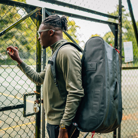 A person with braided hair stands next to a chain-link fence, wearing a green jacket and sunglasses, carrying a large black Racquet Bag Pro from Cancha.