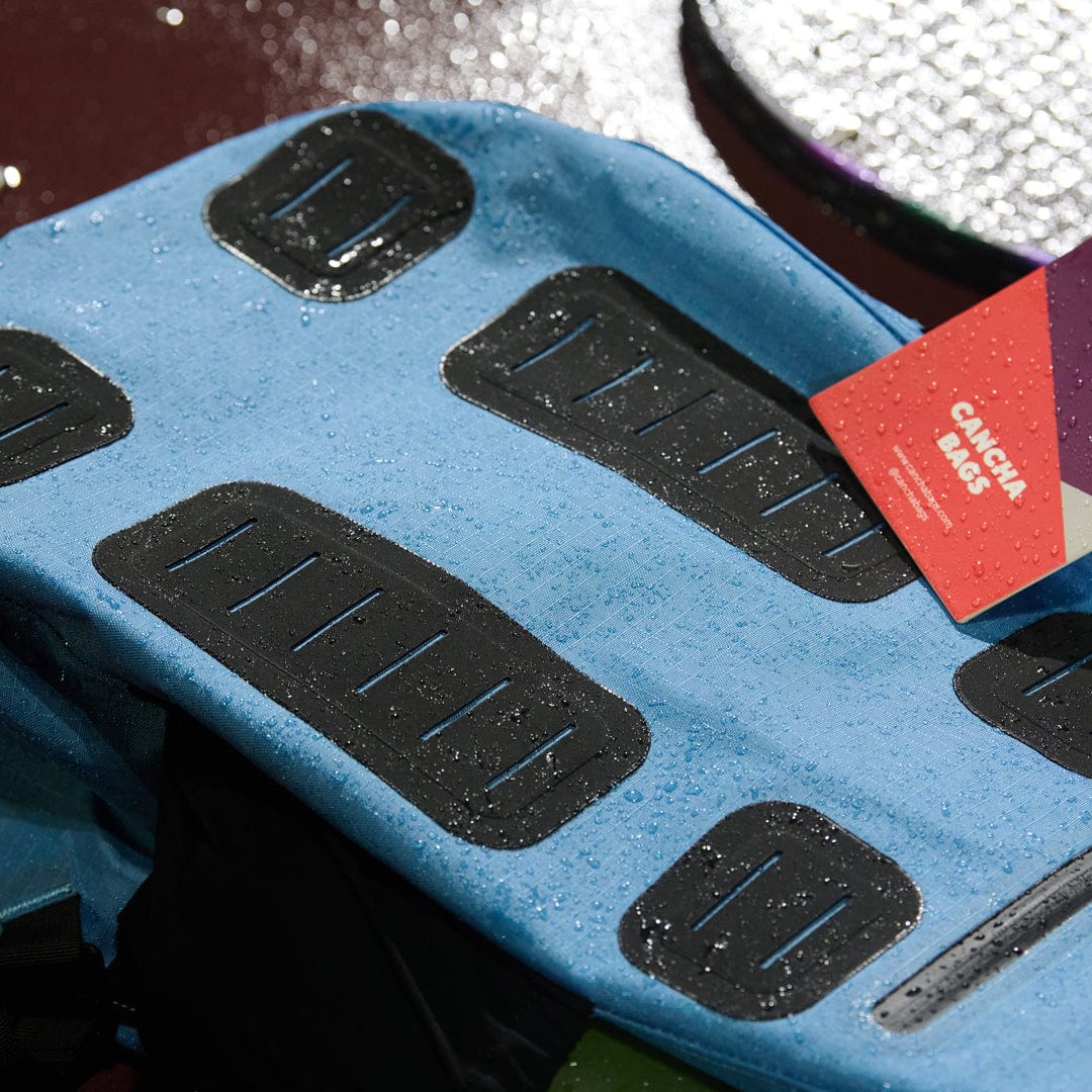 A blue waterproof backpack with black accents and a red tag labeled "Cancha" sits on a wet surface, perfect for those seeking reliable travel accessories from the Tennis Travel Bundle.