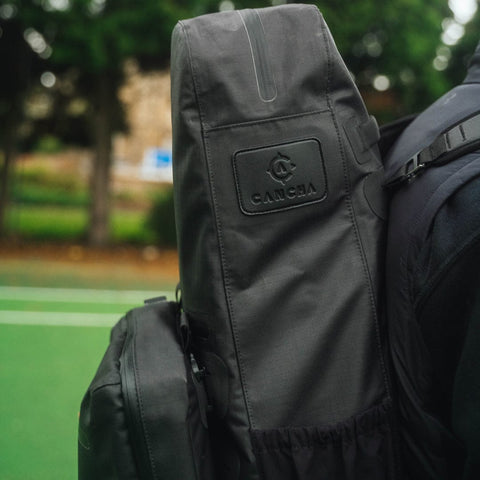 Close-up of a person wearing the black Tennis Travel Bundle backpack by Cancha, featuring a visible "CANCHA" logo, set against a green outdoor surface with trees in the background—ideal for those seeking versatile travel accessories.