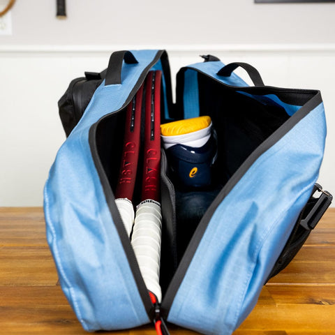 Open the Tennis Travel Bundle from Cancha, featuring a blue Racquet Bag Voyager that holds two tennis rackets, a pair of white and blue athletic shoes with yellow soles, and plenty of space for your travel accessories.