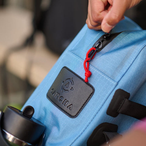 A person unzips a blue Cancha Tennis Travel Bundle backpack featuring a black and red zipper pull. A black water bottle is placed in a side pocket, along with other travel accessories.