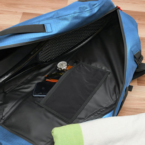 Unzip the blue Cancha Tennis Travel Bundle on a wooden floor, revealing travel accessories such as a racket, watch, sunglasses, and a phone tucked neatly within an internal pocket.