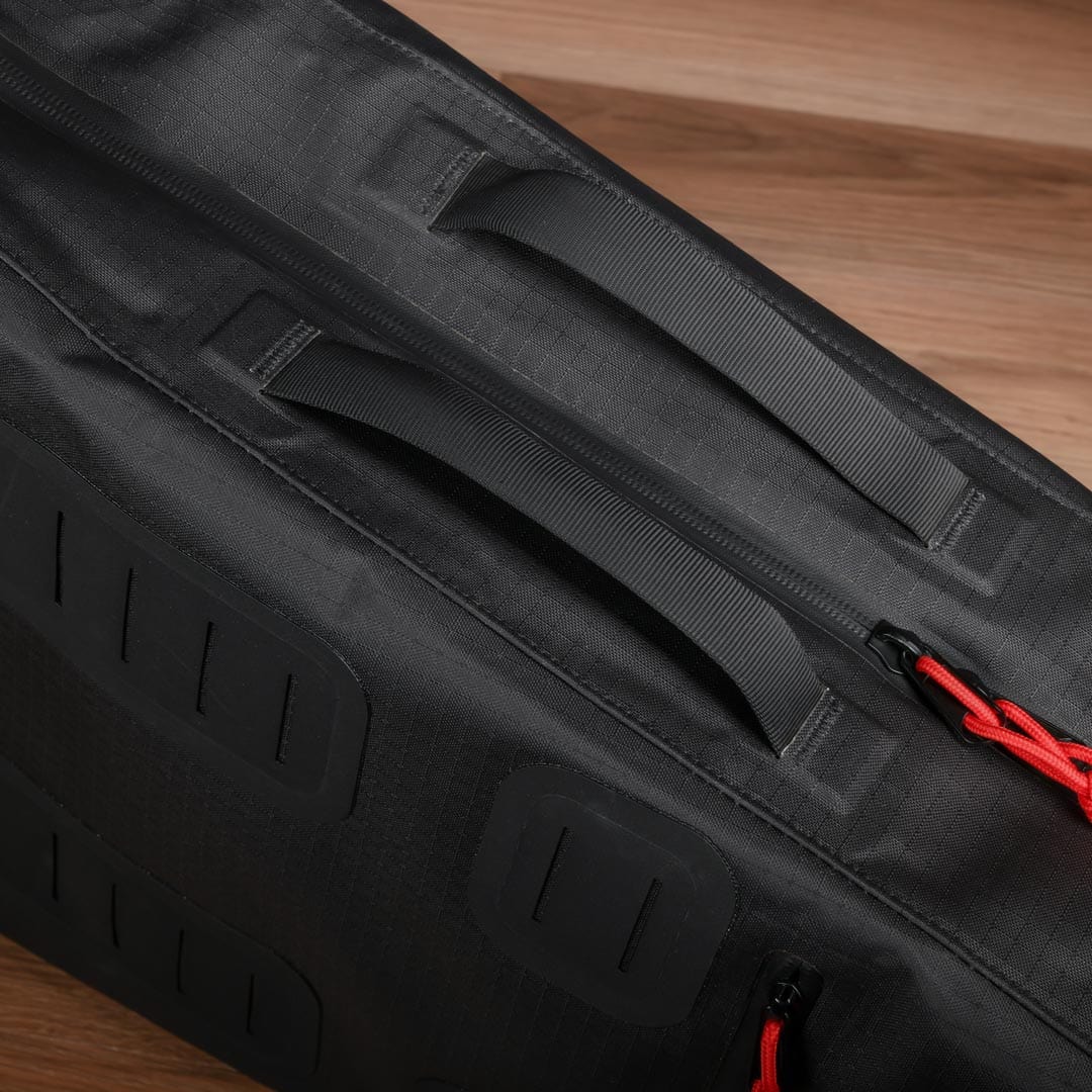 A close-up of the Cancha Tennis Travel Bundle, a black fabric travel accessory featuring a handle, red zipper pull, and rectangular reinforcement patches, placed on a wooden floor.