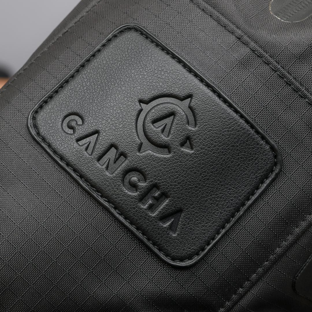 Close-up of the black fabric Tennis Travel Bundle with an embossed "Cancha" logo on a leather patch. Ideal for travel accessories, this bag features wet-dry compartments.