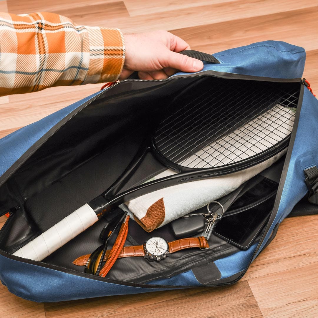 A person’s hand is holding open a Cancha Tennis Travel Bundle, showcasing a tennis racquet, towel, watch, keys, and other small personal items on a wooden floor.