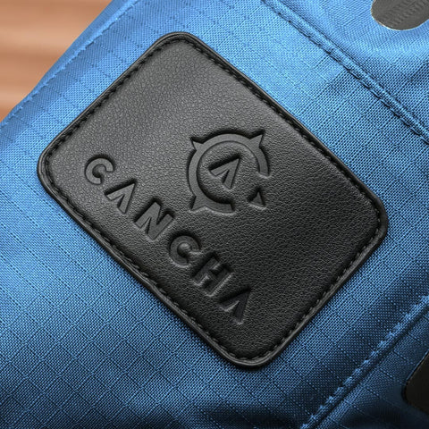 Close-up of a blue fabric item, possibly the Tennis Travel Bundle, featuring a black leather patch with the brand name "Cancha" and a compass logo embossed on it—perfect for travel accessories.