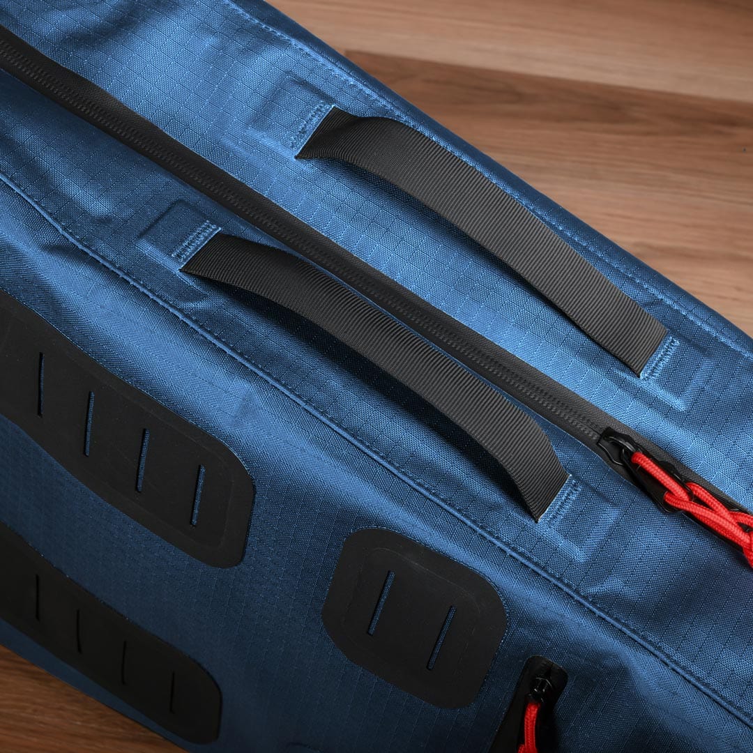 Close-up of a blue fabric Tennis Travel Bundle by Cancha, featuring black handles, zippers, and red zipper pulls, against a wooden background. This versatile travel accessory is perfect for your next adventure.